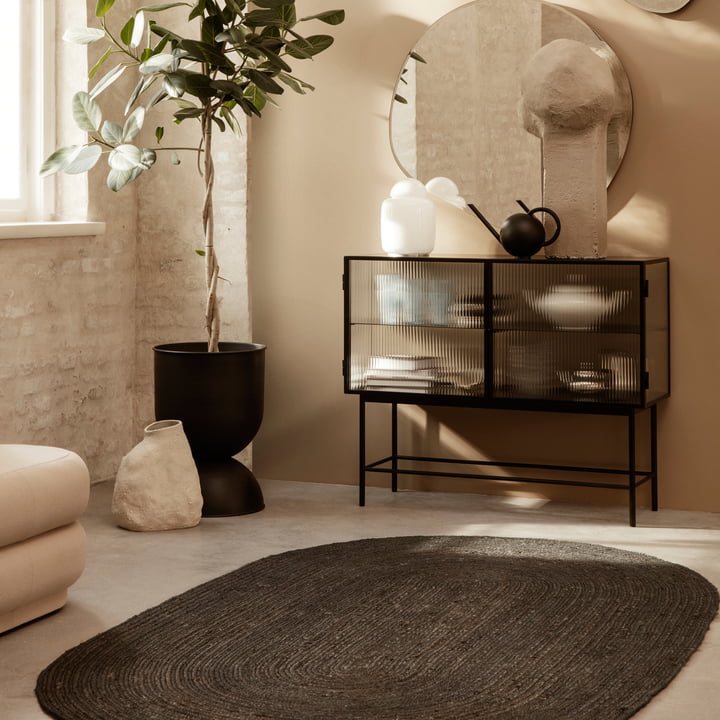 The Eternal Oval Jute Carpet by ferm Living in front of a sideboard and next to a tall houseplant