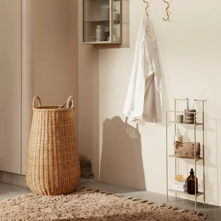 The Braided rattan laundry basket by Ferm Living on a beige carpet next to a small shelf with bathroom accessories