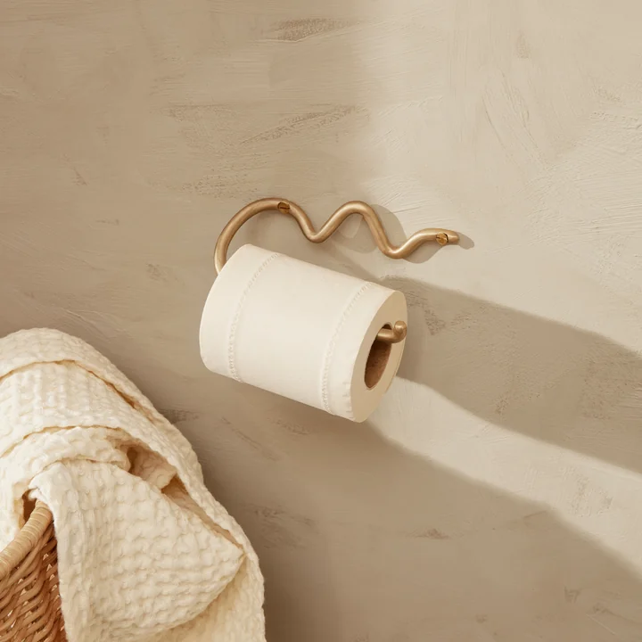 The Curvature toilet paper holder by Ferm Living on a light stone wall next to a rattan basket