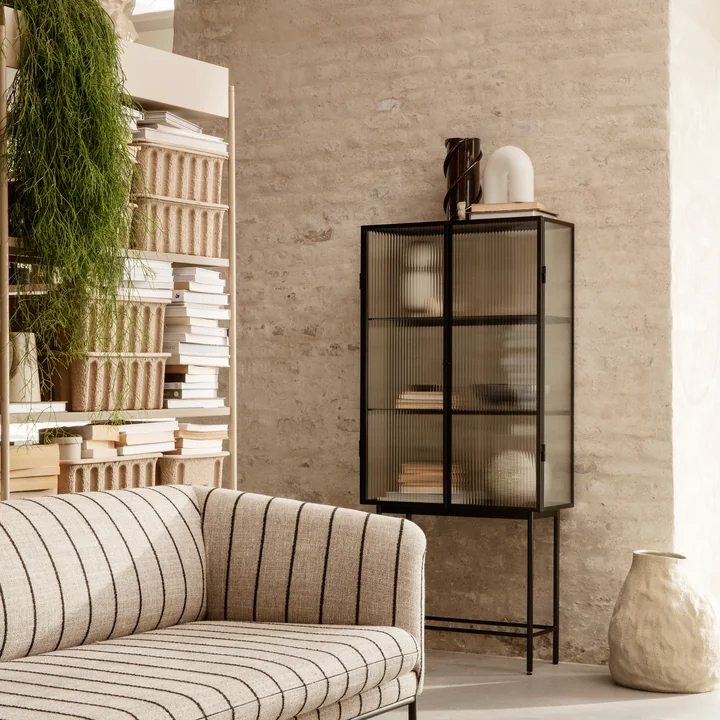 The Haze Display case by Ferm Living with many art vases and books on a stone wall in the living room