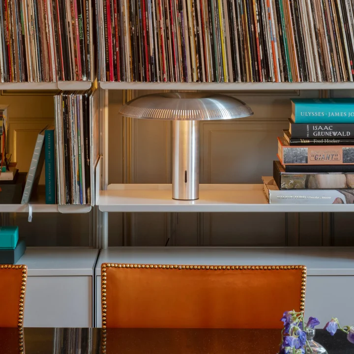 The w203 Ilumina LED table lamp from Wästberg in aluminium on a white shelf between books and exercise books.