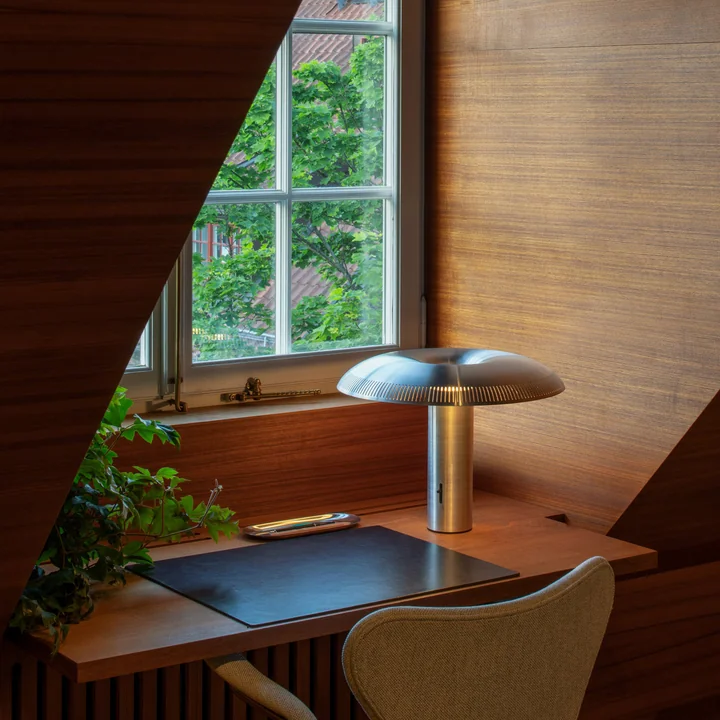 The w203 Ilumina LED table lamp from Wästberg in aluminium on a wooden desk in front of the window