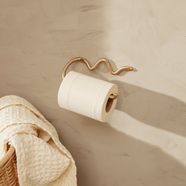 The Curvature toilet paper holder by ferm Living on a light stone wall next to a rattan basket